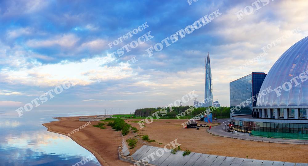 Фотография современного городского пейзажа Санкт-Петербург, Россия. На изображении показано побережье Финский залив с песчаным пляжем и видом на Лахта-центр — знаковый объект современной архитектуры Петербурга. Фотоснимок демонстрирует сферическое здание и прибрежную зону Пляж 300-летия Санкт-Петербурга, подчёркивая сочетание природы и урбанистики. Это изображение передаёт атмосферу современного мегаполиса, где пляжный отдых соседствует с инновационной архитектурой. Иллюстрация может использоваться как картинка или слайд, посвящённые городам России и развитию прибрежных пространств.